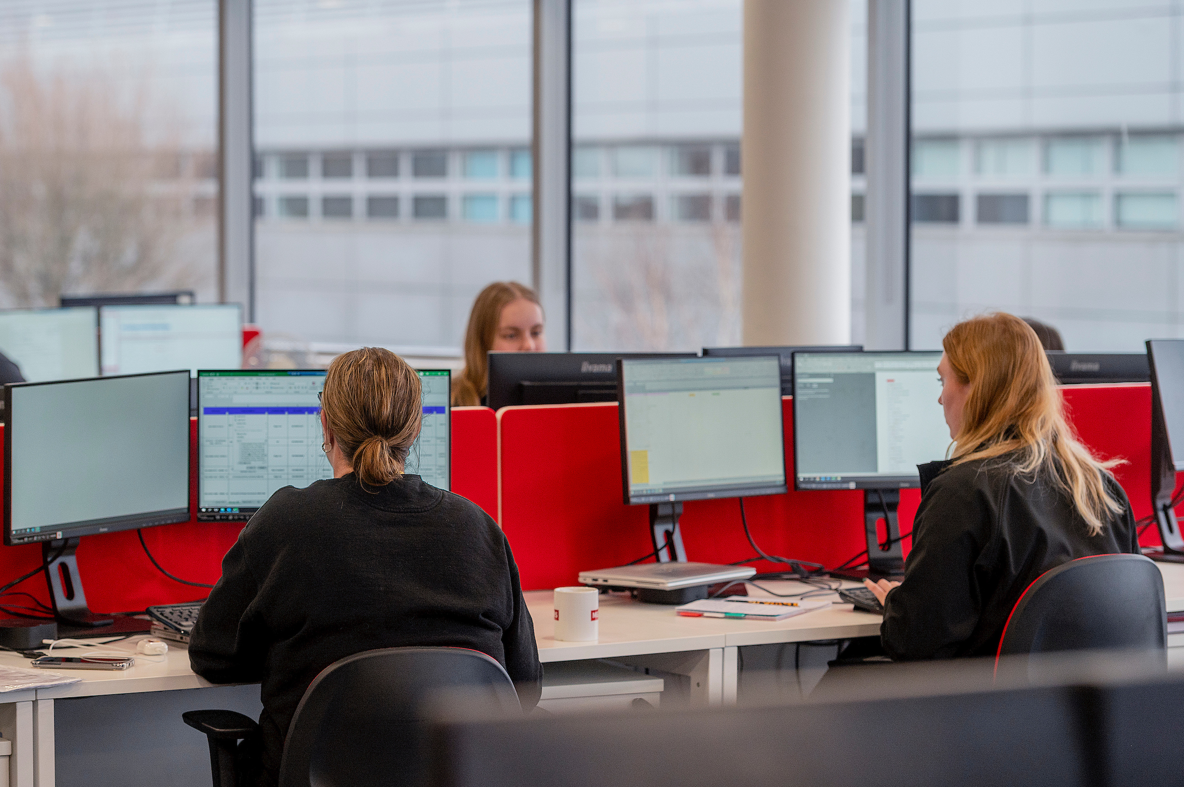 A team of three women work at a modern office space with dual monitors.