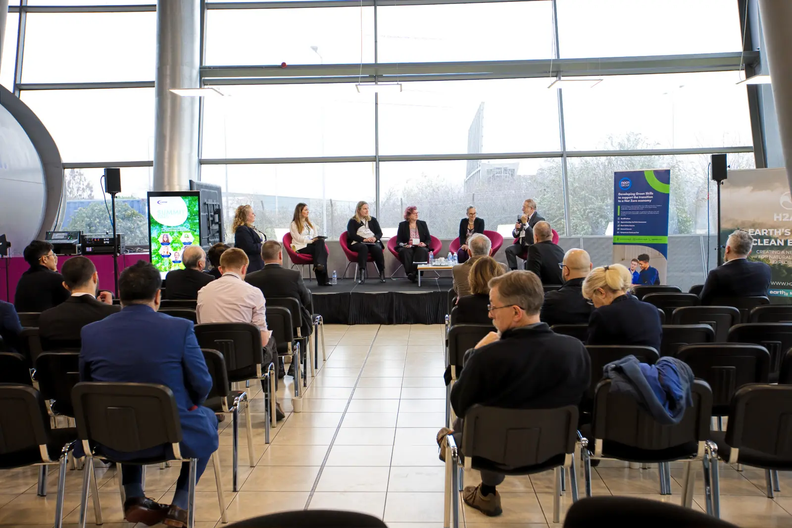 A wide shot of a conference panel discussion taking place in a brightly lit room with large windows. Five speakers (mostly women) are seated on stage facing an audience seated in rows of chairs. A digital screen on the right displays text about "Developing Green Skills" for the Hydrogen economy.