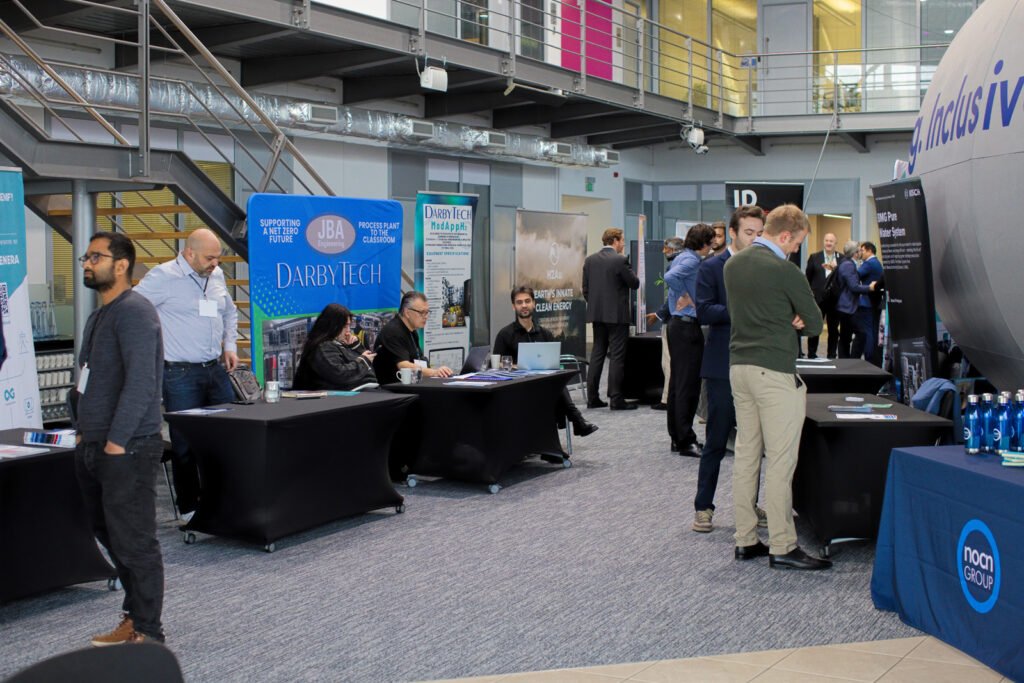 An indoor view of the exhibition and networking area at the Hydrogen Summit. Several small exhibition tables are set up with delegates speaking to company representatives. A large blue banner for "Darby Tech" is visible, as well as a partial view of the large, light-coloured, spherical structure visible in other images.