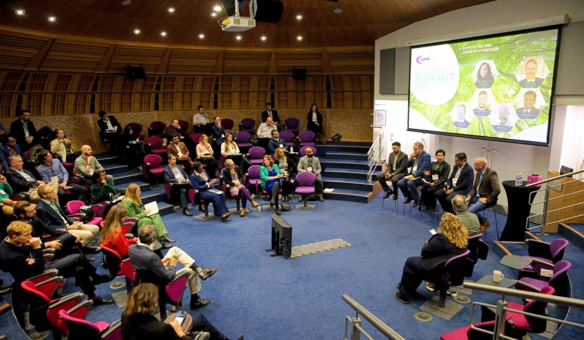 Hydrogen Summit 2025 025 A wide shot of the Hydrogen Summit plenary session taking place in a circular auditorium with tiered seating. A panel of speakers sits in the center facing the audience. A large projection screen shows the summit title and images of speakers.