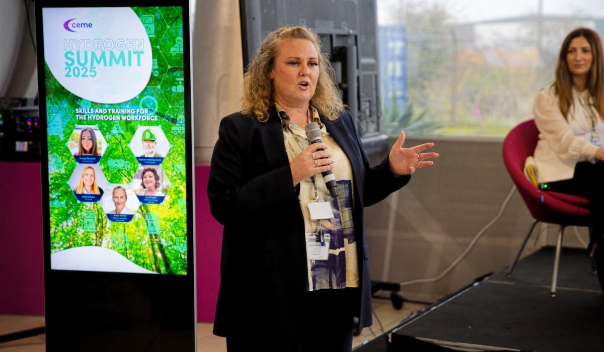 Hydrogen Summit 2025 036 A woman with curly blonde hair wearing a dark jacket speaks into a microphone on a stage. Beside her is a large vertical digital screen displaying the "Hydrogen Summit 2025" title and images of various speakers under the heading "Skills and Training for the Hydrogen Workforce."