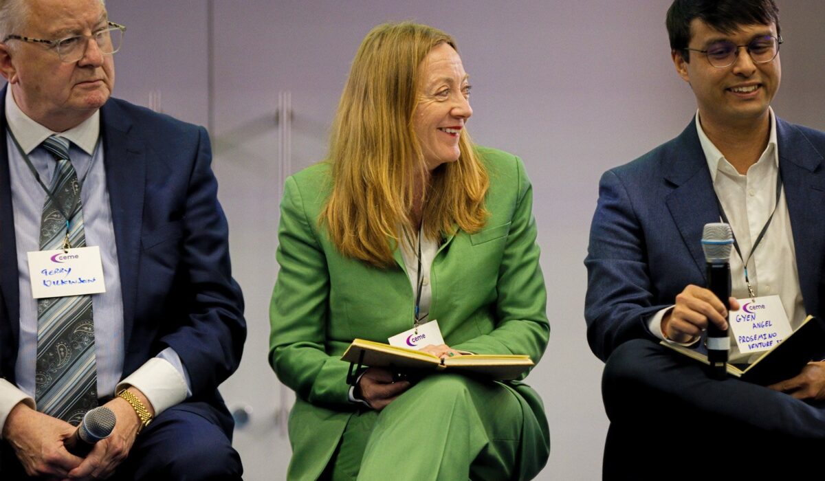 Hydrogen Summit 2025 052 A close-up of three people sitting on a panel stage. A man in a blue suit is on the left, a woman in a bright green suit is smiling and holding a notebook in the center, and a man in a blue suit is smiling and holding a microphone on the right. Name tags are visible.