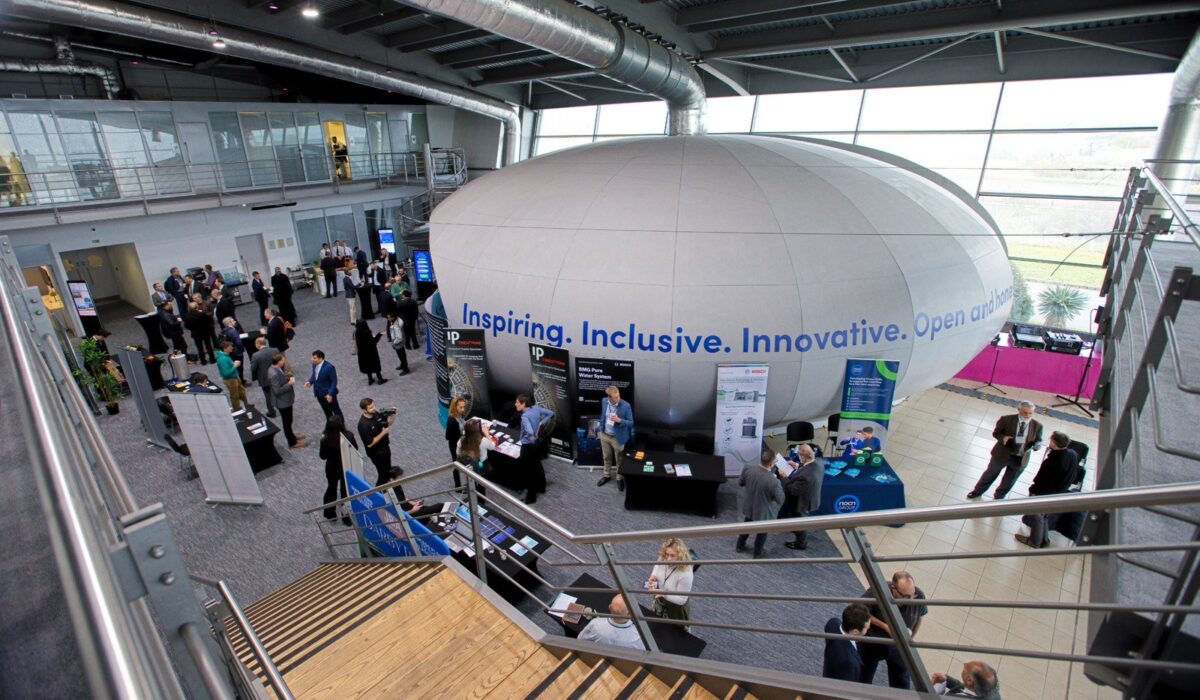 Hydrogen Summit 2025 132 Overview of a large, modern indoor venue for the Hydrogen Summit. Attendees network around tables near a large, central, egg-shaped structure with the text, "Inspiring. Inclusive. Innovative. Open and Honest." The image is taken from an upper level looking down wooden stairs.