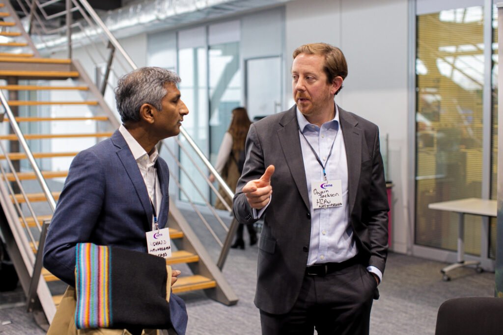 Two men in business attire are engaged in conversation in a modern interior setting near a staircase. The man on the right is gesturing with his finger as he speaks. Both wear CEME event badges.