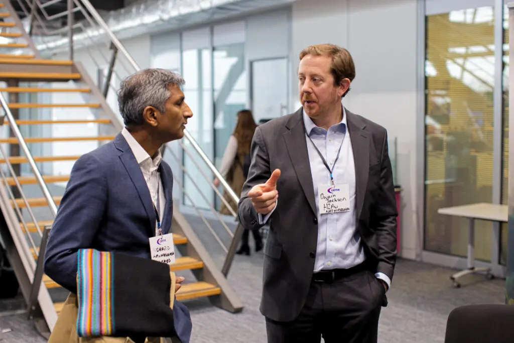 Two men in business attire are engaged in conversation in a modern interior setting near a staircase. The man on the right is gesturing with his finger as he speaks. Both wear CEME event badges.