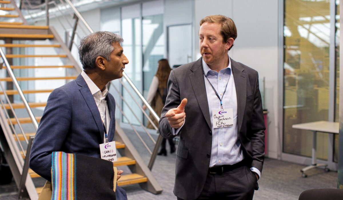 Hydrogen Summit 2025 142 Two men in business attire are engaged in conversation in a modern interior setting near a staircase. The man on the right is gesturing with his finger as he speaks. Both wear CEME event badges.