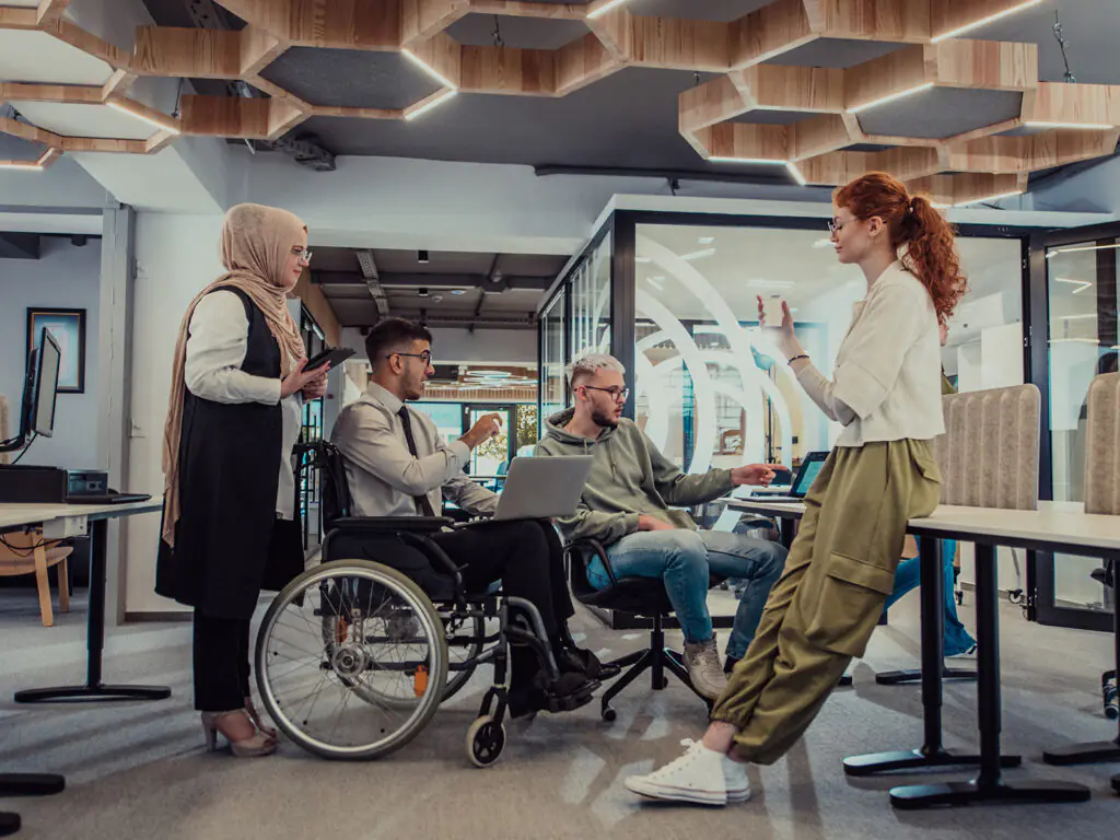 A four person project team gather round a desk in discussion. Two women are standing, and one man is in a wheelchair while his colleague sits beside him controlling a computer.